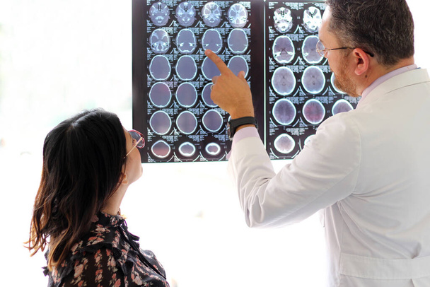 Lekarz medycyny specjalizujący się w neurologii wyjaśnia tomografię mózgu latynoskiej kobiecie w okularach w gabinecie. - Zdjęcie, obraz
