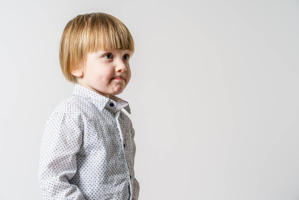 Half length portrait of small blonde caucasian boy standing in front of white wall looking to the side - little child studio portrait with copy space - Foto, Imagen