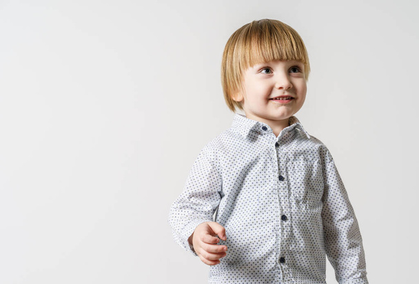 Halbes Porträt eines kleinen blonden kaukasischen Jungen, der vor einer weißen Wand steht und zur Seite lächelt - kleines Kinderatelier-Porträt mit Kopierraum - Foto, Bild