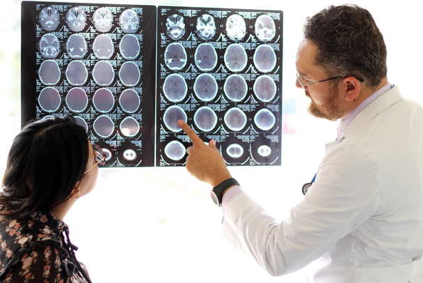 Médico especialista en Neurología explica tomografía cerebral de rayos X a una mujer latina con gafas en su consultorio - Foto, Imagen