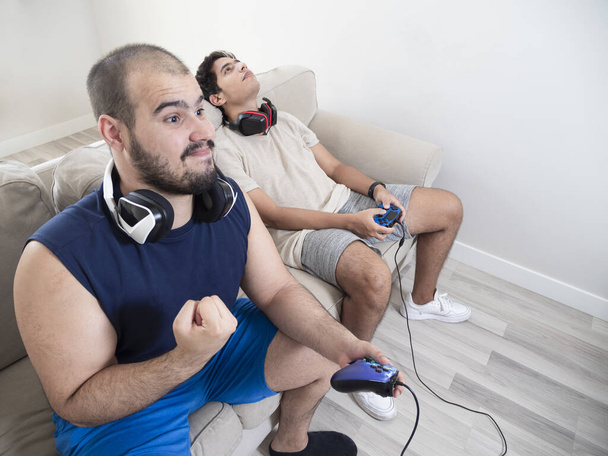 Dwóch młodych ludzi grających w gry wideo z kontrolami gier wideo na białej sofie w białym pokoju, białej młodzieży i ciemnoskórej. - Zdjęcie, obraz