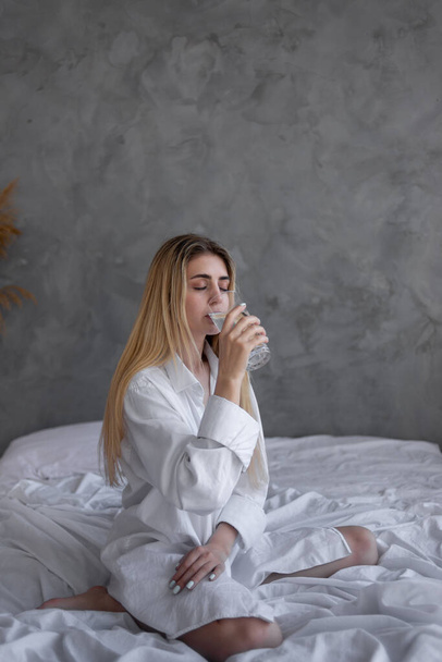 Egy pohár tiszta terv egy fiatal nő kezében. Nő fehér ing ül az ágyban, reggel este étel és italok, egészséges életmód - Fotó, kép