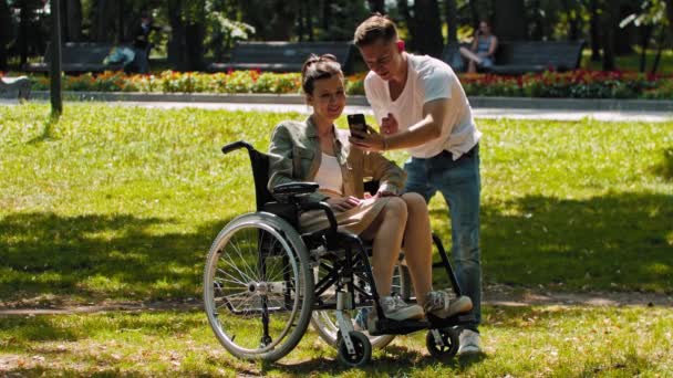 Tekerlekli sandalyedeki kadın ve arkadaşı yaz parkında vakit geçiriyorlar ve telefonda selfie çekiyorlar. - Video, Çekim