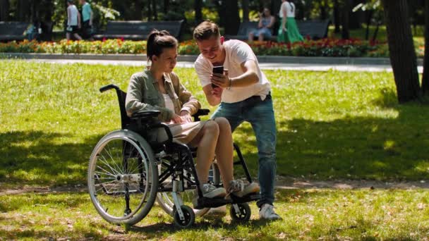 Mujer en silla de ruedas y su amiga pasando tiempo en el parque de verano y el hombre mostrando sus fotos desde su teléfono - Metraje, vídeo