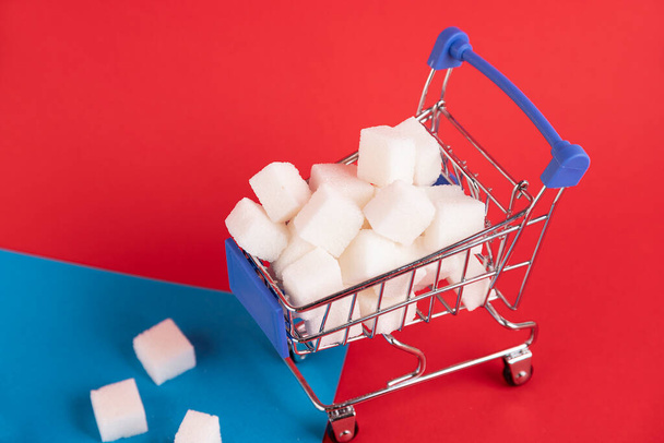 A shopper's cart is filled with refined sugar cubes on a red background. Copy space. - Photo, image