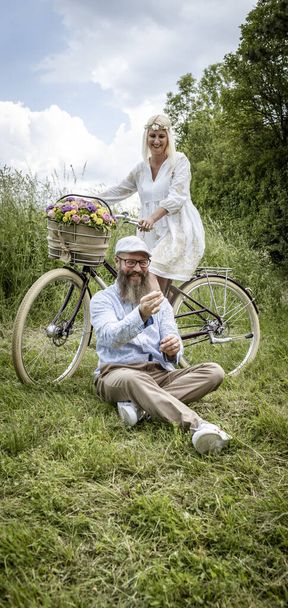donna bionda in abito bianco e suo marito in posa con bicicletta decorato fiori cesto in natura  - Foto, immagini
