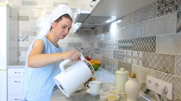 Kobieta po prysznicu z ręcznikiem na głowie przygotowuje kawę w kuchni. Wlać wrzącą wodę z czajnika elektrycznego do filiżanki. Przygotowanie śniadania - Materiał filmowy, wideo
