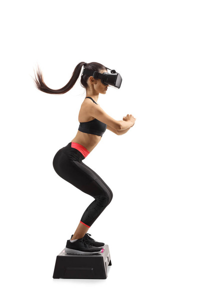Full length profile shot of a young woman exercising on a step aerobic platform with a vr headset isolated on white background - Φωτογραφία, εικόνα