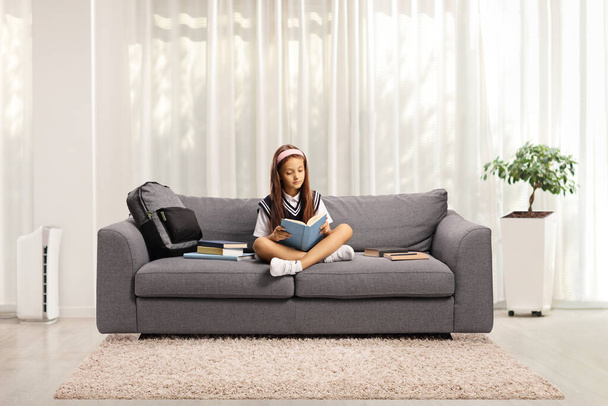 Schoolgirl in a uniform sitting on a couch and reading a book at home in a living room - Foto, afbeelding