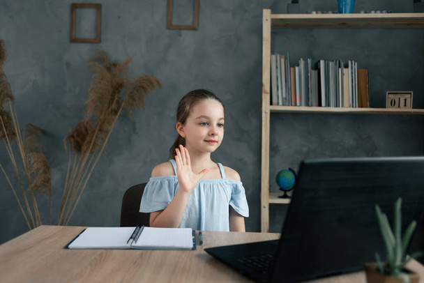 Little Caucasian girl smiling and waving her hand while sitting at home at the table with a laptop. Online communication, distance learning. Copy space. - Photo, Image