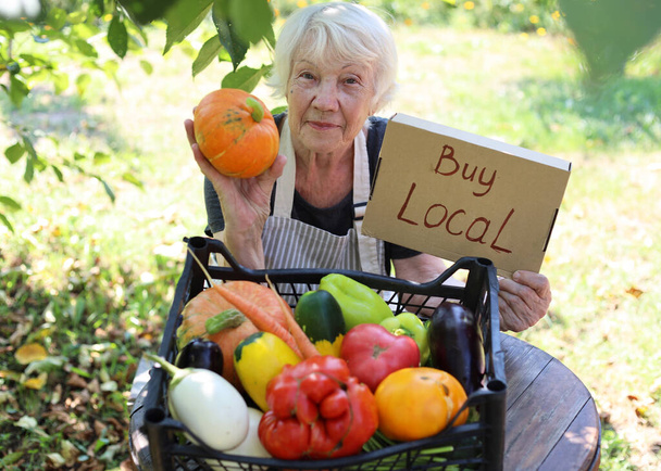 高齢者の白髪の女性が地元のファーマーズマーケットで新鮮な旬の野菜を販売しています。地元の農産物を買う - 写真・画像