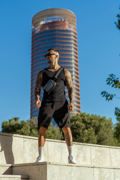 A vertical shot of a Caucasian sporty male with tattoos on a background of a high-rise building - Foto, Imagen