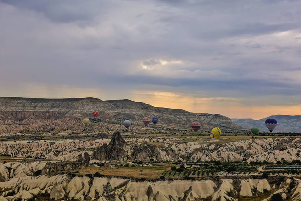 Dawn over Cappadocië. De bewolkte lucht is oranje gemarkeerd. Kleurrijke ballonnen vliegen over verbazingwekkende rotsen met gevouwen hellingen. Een berg met een platte top in de verte. - Foto, afbeelding