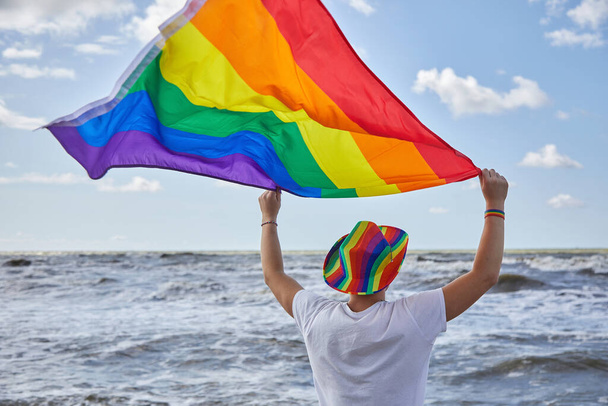 Man standing by the sea and holding the LGBTQ pride flag. LGBTQ concept - Foto, imagen