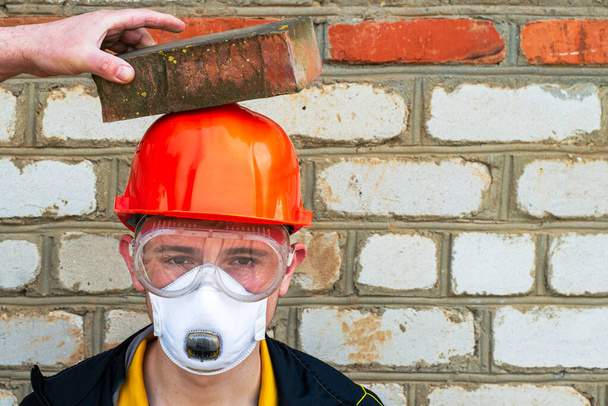 een bouwvakker in een beschermende bouwhelm, een masker en een veiligheidsbril tegen de achtergrond van een bakstenen muur, een helm beschermt tegen een baksteen impact copyspace. Hoge kwaliteit foto - Foto, afbeelding