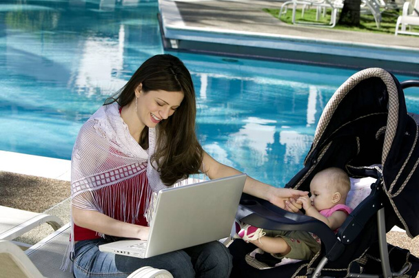 Äiti käyttää kannettavaa tietokonetta tyttärensä vieressä uima-altaalla
 - Valokuva, kuva
