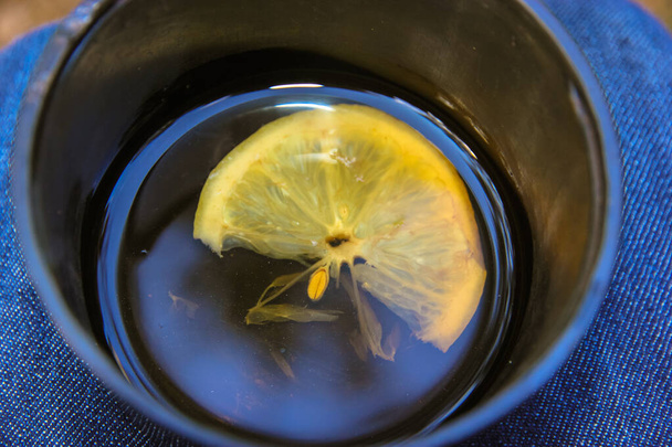 Fincanın üstünde bir dilim limonlu siyah çay. Termos bardağının içinde güzel kokulu sıcak bir içecek yatıyordu. Soğuk algınlığı sırasında kullanışlı bir içecek mevsimsel hastalığın ilk belirtileri, grip. Denim arkaplanı. - Fotoğraf, Görsel