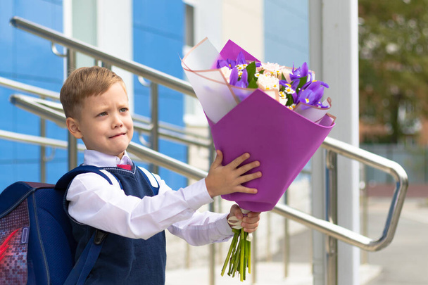 Un lindo niño de primer grado en un uniforme escolar con un hermoso ramo de flores de colores en el patio de la escuela después de las vacaciones del 1 de septiembre. Día del conocimiento. Enfoque selectivo - Foto, imagen