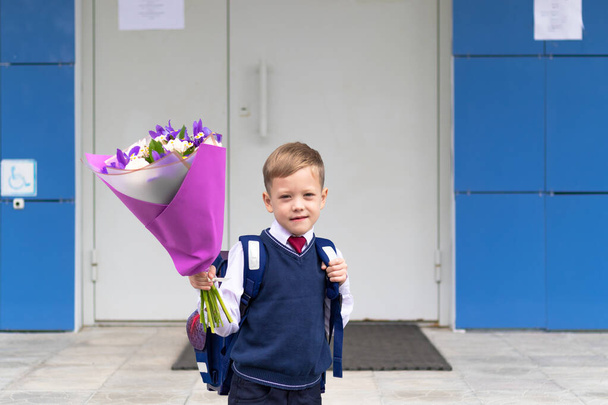 Ein süßer Erstklässler in Schuluniform mit einem schönen Strauß bunter Blumen auf dem Schulhof nach den Ferien am 1. September. Wissenstag. Selektiver Fokus - Foto, Bild