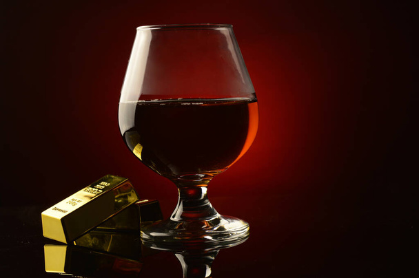 A concept of wealth utilizing a couple gold bullion bars next to a rich Cognac drink over a dark reflective red and black background. - Photo, image