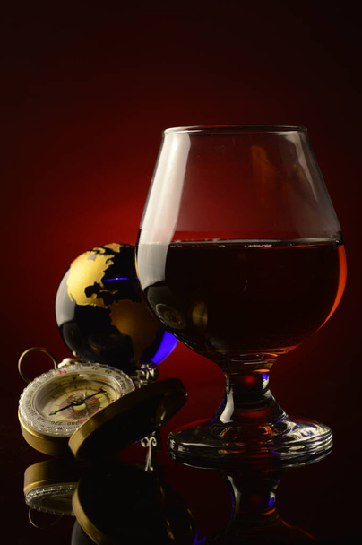 A glass of Cognac with a compass and globe over a dark red and black background. - Photo, image