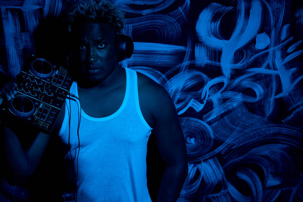 Confident black man in a white T-shirt holds a DJ console in his hands and listens to music with black headphones against the background of graffiti. Professional African-American DJ  - Foto, afbeelding