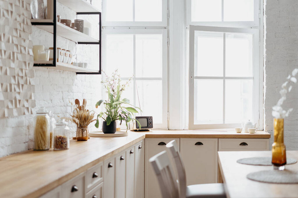 Εσωτερικό από λευκή κουζίνα με ξύλινα μοντέρνα έπιπλα. Κλείσιμο του καθαρού σκανδιναβικού σχεδιασμού στο σπίτι, επιτραπέζιος μετρητής στο φως. Γυάλινη κουζίνα στο τραπέζι, δωμάτιο με ρετρό διακόσμηση. - Φωτογραφία, εικόνα