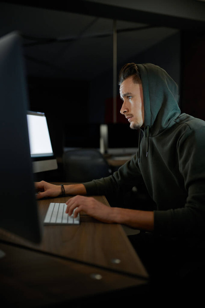 Young male internet hacker in hood sitting at monitors. Illegal web programmer at workplace, criminal occupation. Data hacking, cyber security - Fotó, kép