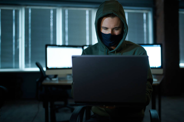 Male internet hacker in hood holds laptop, front view. Illegal web programmer at workplace, criminal occupation. Data hacking, cyber security - Foto, Imagen