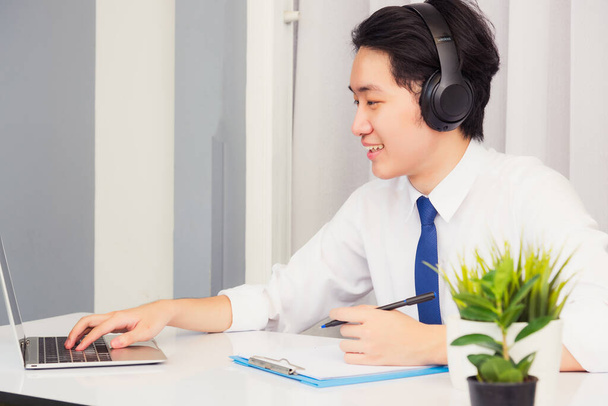 Work from home, Asian young businessman wear suit video conference call or facetime he smiling sitting on desk using laptop computer and write paper notebook information at home office - Photo, image