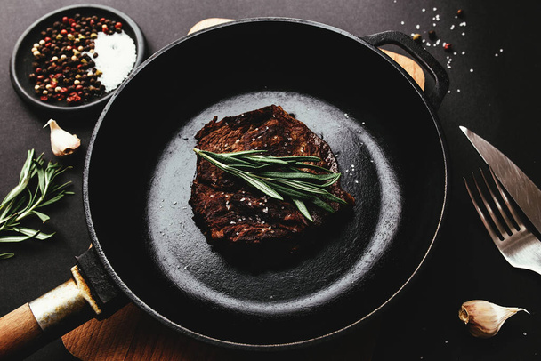 Cooked ribeye steak on cast-iron pan and board with pepper, rosemary, salt, garlic, knife, fork, butter on black background - Foto, immagini