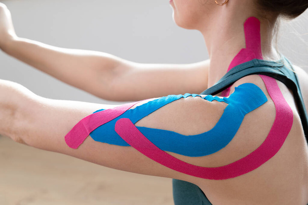 Closeup of a young fit womens shoulder with elastic therapeutic kinetic tape on her shoulder. Performing exercise at home. Kinesiology physical therapy. - Φωτογραφία, εικόνα