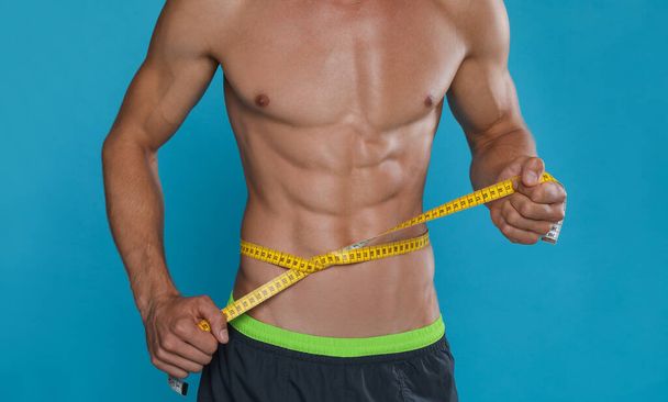 Shirtless man with slim body and measuring tape around his waist on light blue background, closeup - Foto, immagini