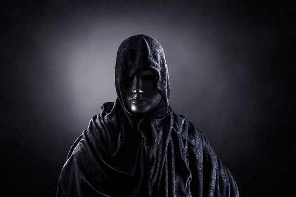 scary halloween concept. man in black hooded hoodie with hood and looking at camera on dark background - Fotó, kép