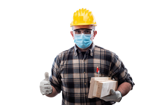 Carpenter worker isolated on white background wears surgical mask to prevent Coronavirus spread, makes OK sign with thumb up. Preventing Pandemic Covid-19 at the workplace. - Fotoğraf, Görsel