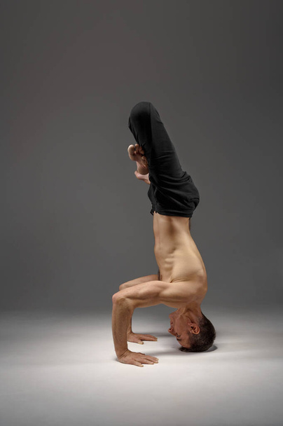 Male yoga standing on his head and hands, meditation, grey background. Strong man doing yogi exercise, asana training, top concentration - Photo, image