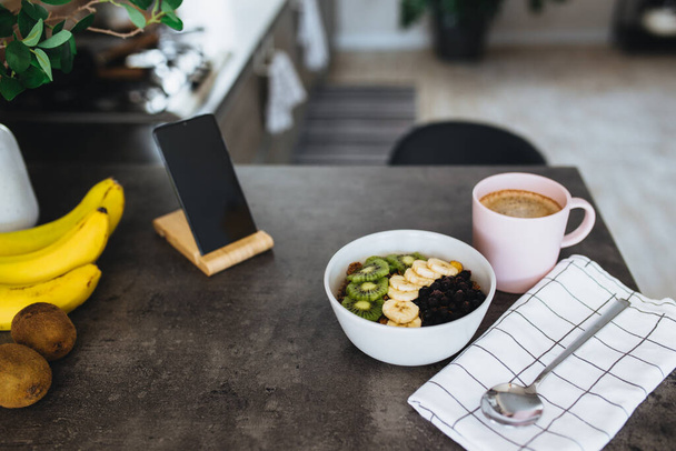 Pink coffee cup, bowl with chopped tropical fruits kiwi and banana, blueberries, spoon and mobile phone on bar counter in stylish loft kitchen. Blurred background. High quality photo - Photo, Image