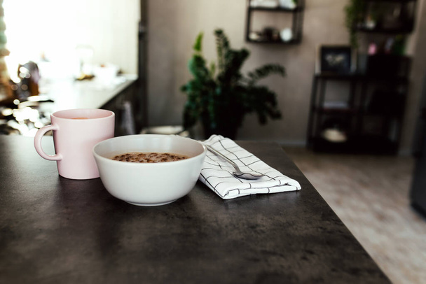 Xícara de café rosa, tigela com frutas tropicais picadas kiwi e banana, mirtilos, colher na toalha no balcão de bar na elegante cozinha loft. Fundo desfocado. Foto de alta qualidade - Foto, Imagem