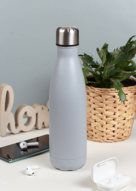 Grey insulated bottle on white desk surrounded by modern gadgets and plant front view - Φωτογραφία, εικόνα