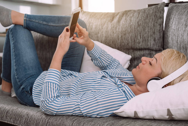 Happy woman with headphones and mobile phone listening to music and relaxing on sofa at cozy home - Foto, Imagen
