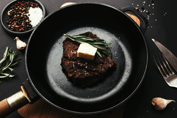 Cooked ribeye steak on cast-iron pan and board with pepper, rosemary, salt, garlic, knife, fork, butter on black background - Φωτογραφία, εικόνα