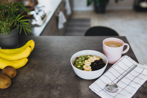 Pink coffee cup, bowl with chopped tropical fruits kiwi and banana, blueberries, spoon on bar counter in stylish loft kitchen. Blurred background. High quality photo - Foto, Imagem