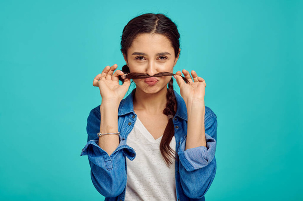 Funny young woman shows a mustache, blue background, positive emotion. Face expression, female person looking on camera in studio, emotional concept, feelings - Фото, изображение