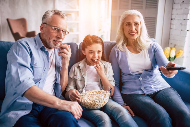  Счастливая девочка, бабушка и дедушка смотрят телевизор с попкорном - Фото, изображение