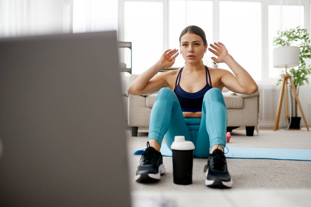 Slim woman sits on mat, online fitness training at the laptop. Female person in sportswear, internet sport workout, room interior on background - Valokuva, kuva