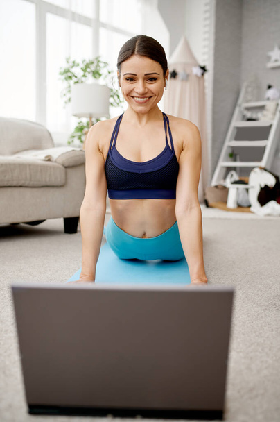 Χαμογελαστή κοπέλα κάθεται στο πάτωμα στο σπίτι, σε απευθείας σύνδεση προπόνηση ταιριάζει στο φορητό υπολογιστή. Γυναίκα στα αθλητικά είδη, internet sport workout, δωμάτιο εσωτερικό - Φωτογραφία, εικόνα