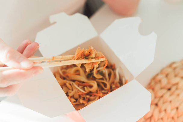 Wok noodles in takeaway box. Woman eating with chopsticks, close up view on female hands. Chinese traditional food with vegetables and seafood. - Photo, Image