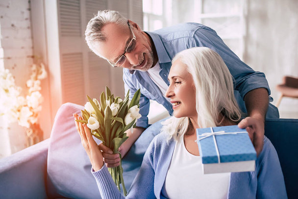De glimlachende oudere man geeft bloemen en een geschenk aan een vrouw - Foto, afbeelding