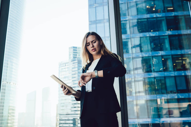 Puzzled female corporate director with modern digital tablet in hand standing near panoramic window in office and checking time on wearable smartwatch feeling confused indoors, business concept - Valokuva, kuva