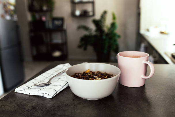 Pink coffee cup, bowl with chopped tropical fruits kiwi and banana, blueberries, spoon spoon on towel on bar counter in stylish loft kitchen. Blurred background. High quality photo - Φωτογραφία, εικόνα